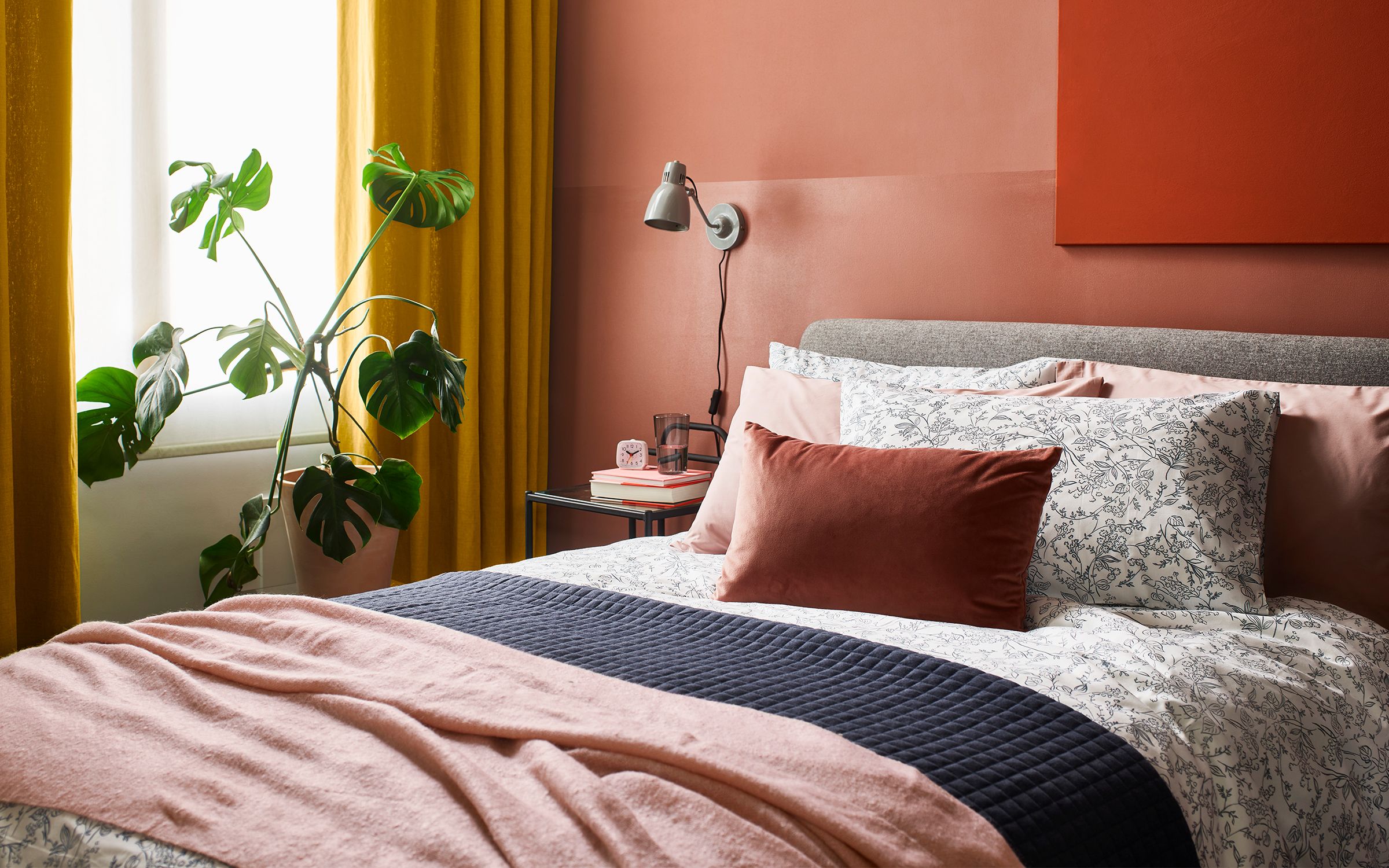 bed with pink and patterned pillows, pink throw, navy throw, mustard curtains, black sidetable, large plant