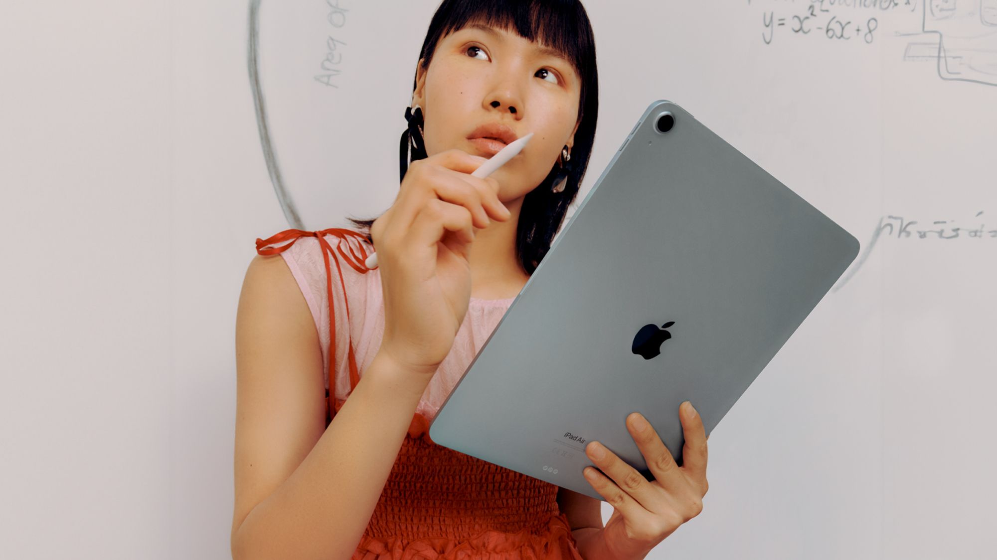 student holding a ipad and apple pencil looking thoughtful while standing against a wall with diagrams and math equations on it