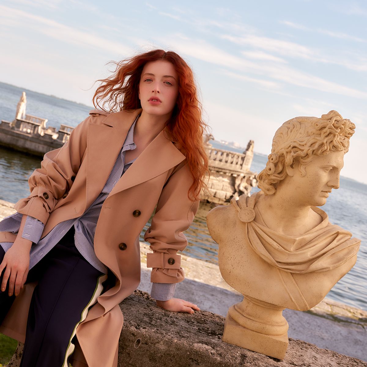 Image of a lady with red hair, sitting on the side of the harbour wearing a trench coat