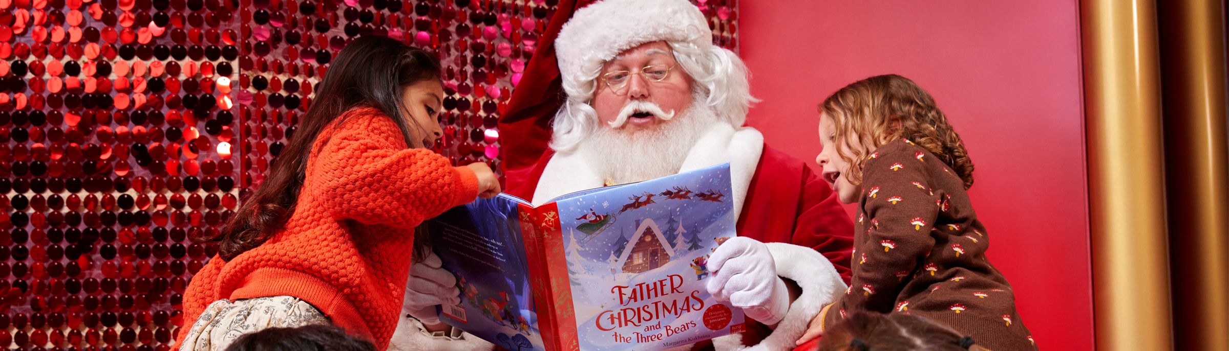 Image of a santa reading a Christmas book to children, with a red sparkly backdrop