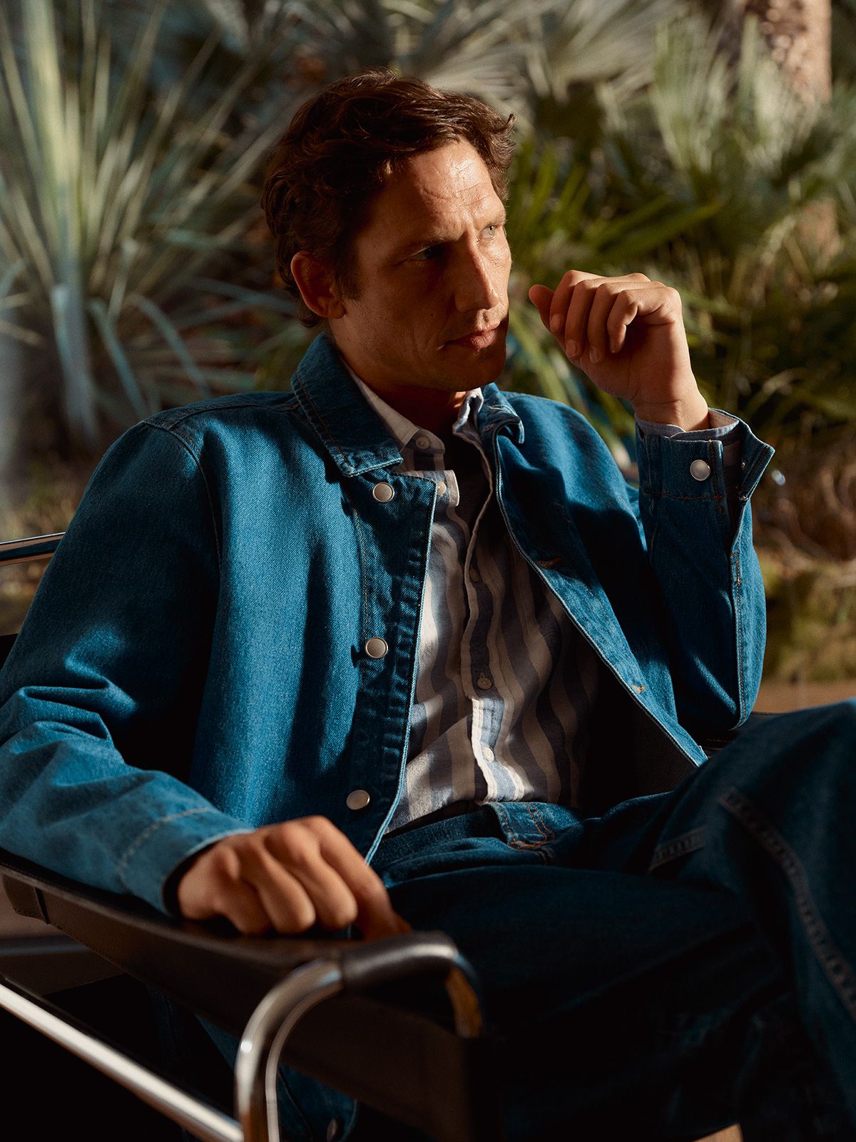 Image of a man sitting down in a denim jacket with a jungle backdrop behind him