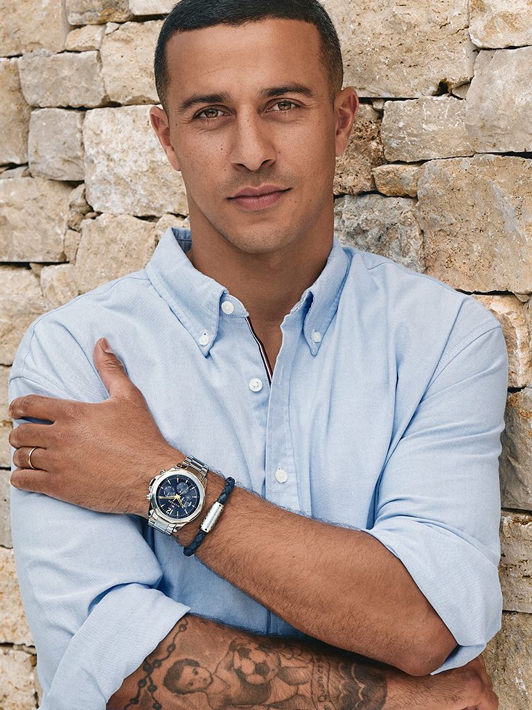 Man in blue shirt stood against a brick wall showing his wrist with a watch on