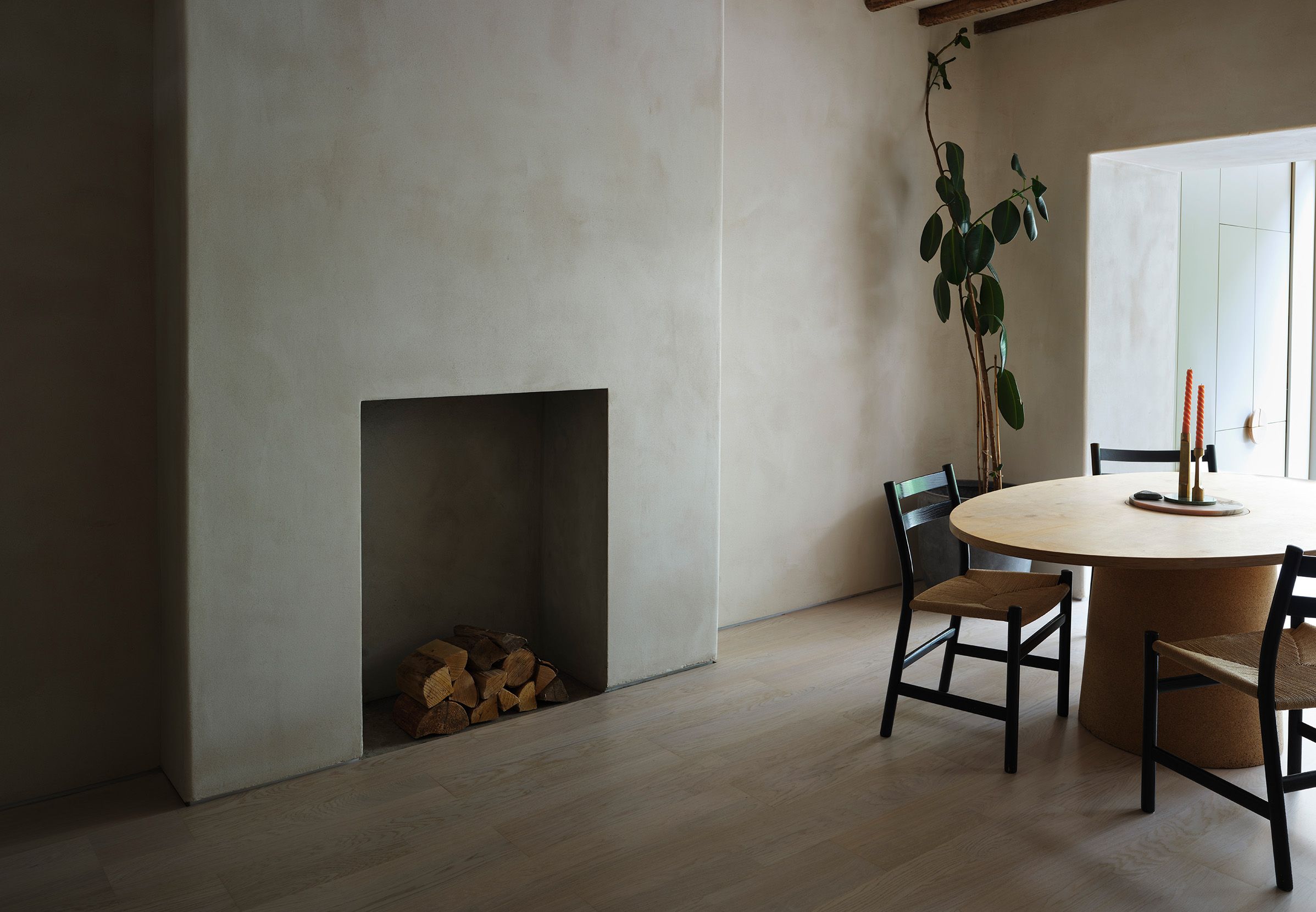 Wood flooring room with plastered wall and modern fire place. Wooden round table with chairs around it. Candles on the table and a leafy plant in the back corner of the room.